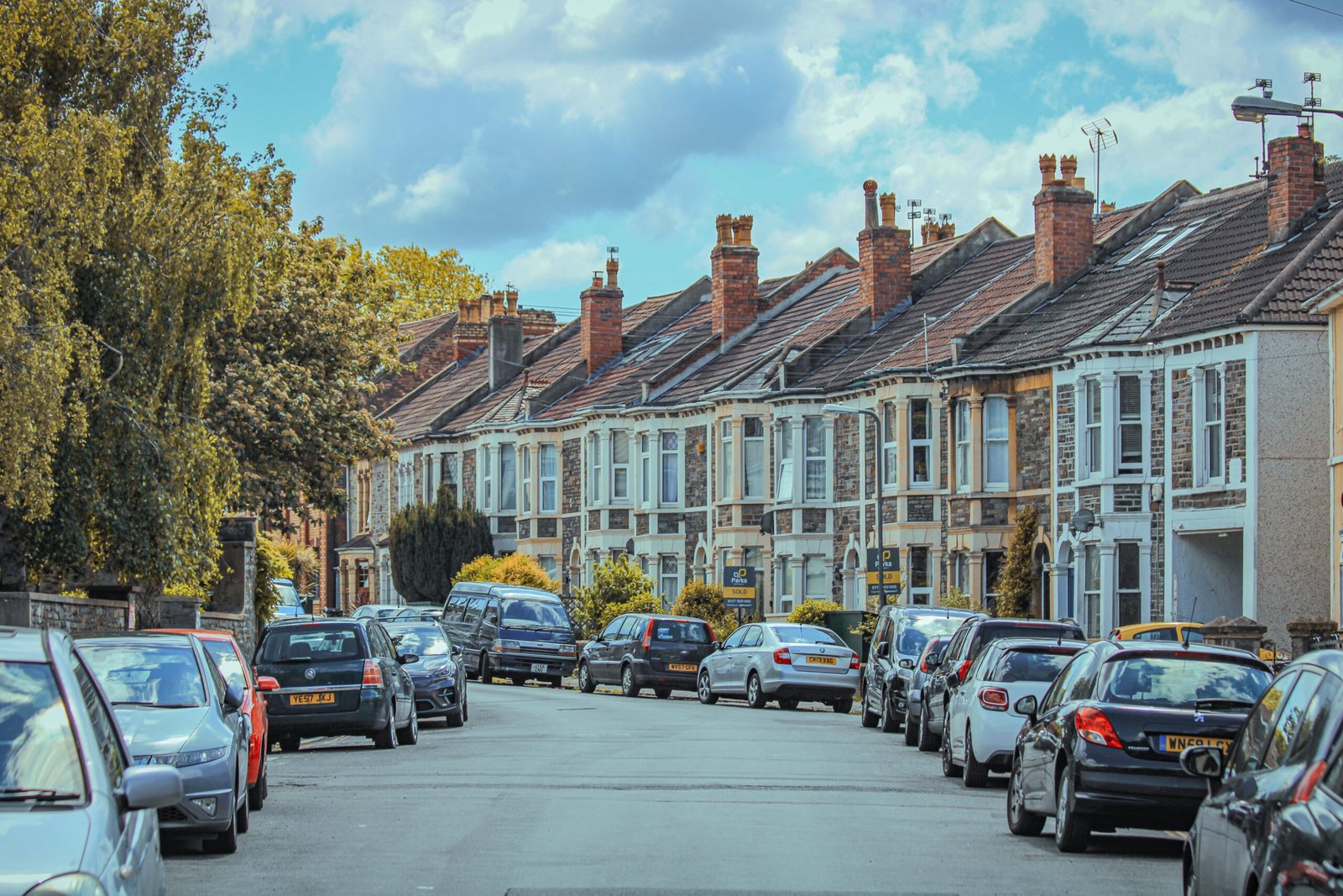 A tree-lined street of British terraced houses with cars parked outside